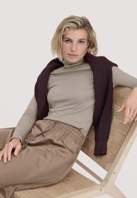 Beiger Langarmrollkragenpullover unter einem gerippten, dunkelburgunderfarbenen Pullover, kombiniert mit lockeren braunen Hosen, sitzend auf einem geflochtenen Stuhl.