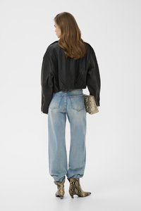 Femme aux longs cheveux bruns portant une veste en cuir noire, un jean bleu clair et des bottes à imprimé serpent, tenant une pochette assortie à l'imprimé serpent.