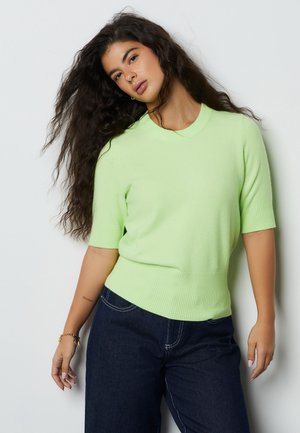 Joven mujer con el pelo largo y rizado, vistiendo un suéter de manga corta verde claro y vaqueros oscuros, posando frente a un fondo claro y sencillo.