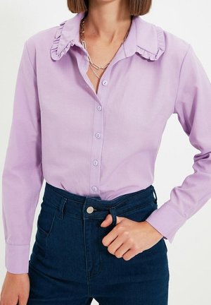 Frau trägt eine hellviolette, geknöpfte Bluse mit Rüschenkragen und dunkle, hoch taillierte Jeans, Hand ruht auf dem Bund.