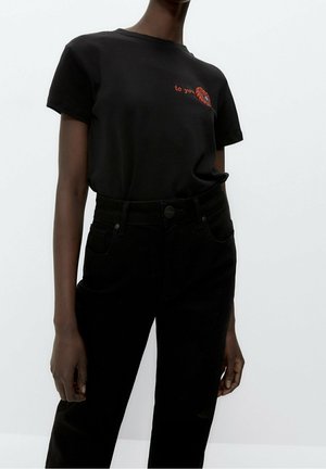 Osoba nosząca dopasowany czarny T-shirt z czerwonym wyszywanym ustem i napisem "to you" na piersi, w połączeniu z czarnymi spodniami z wysokim stanem.