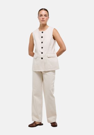 Femme debout portant un gilet sans manches écru avec des boutons noirs, un pantalon écru assorti, et des chaussures plates tissées marron.