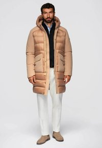 Beige pufferjack met een capuchon, voorzien van grote zakken aan de voorkant en een ritssluiting. Gedirigeerd over een zwarte trui en lichte broek, aangevuld met bruine laarzen.