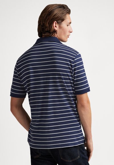 Vīrietis, kas stāv ar muguru, valkājot tumši zilu polo krekli ar baltām horizontālām svītrām un tumšas džinsas uz vienkrāsaina balta fona.