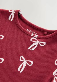 Burgundská textúrovaná tkanina s bielymi vzormi mašlí, s ryhovaným výstrihom. Štítok znie "NEXT." Detailný záber na odev.