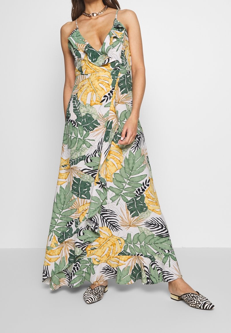 Femme portant une robe longue imprimée de feuilles tropicales vertes, jaunes et blanches, avec fines bretelles et volant à l'avant, associée à des chaussures à motifs noirs et blancs.