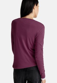 Femme aux longs cheveux bruns portant un tee-shirt long violet et un jean noir, de dos contre un fond uni.