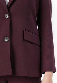 Nærbilde av en person som har på seg en mørk lilla blazer med to knapper og en klaffelomme på høyre side.