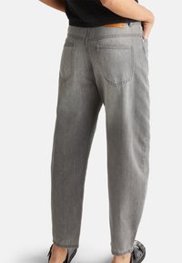 Graue Denim-Hose mit lockerer Passform, ausgestattet mit Gesäßtaschen und einem schwarzen Bund, getragen mit schwarzen Schuhen. Sichtbare Nahtdetails.