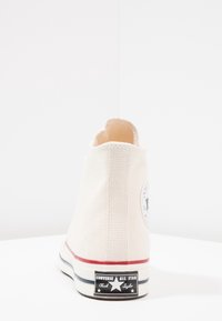 Beige high-top canvas sneaker with rubber sole, featuring a circular logo patch, red accent line, and black label on the heel.