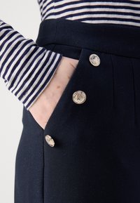 Pantalons bleu marine avec une poche latérale et des accents de boutons argentés, associés à une chemise à manches longues bleu marine et blanche à rayures.