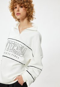 Witte fleece sweatshirt met zwarte accenten, halve rits kraag en een grafische print met de tekst "MONTECARLO ATHLETIC VIBES" over de voorkant.