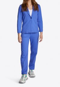 Eine Frau trägt ein hellblaues Blazer und passende Hose, kombiniert mit weißen und grünen Sportsneakers, und steht vor einem weißen Hintergrund.