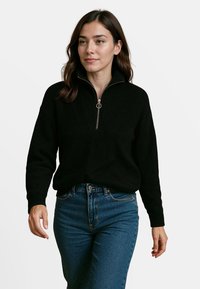 Zwarte gebreide pullover met een kwart ritssluiting en ribbelstructuur; lange mouwen en een relaxed fit, gecombineerd met high-waisted blauwe jeans.