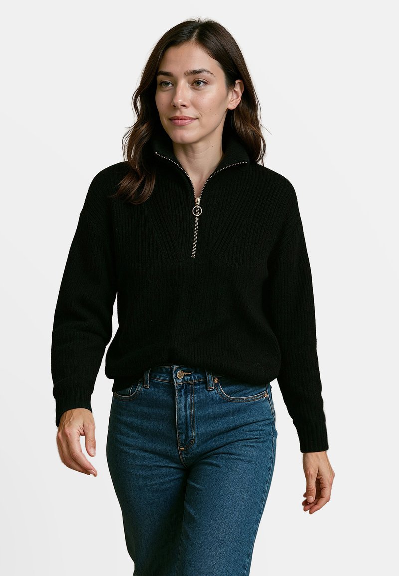 Zwarte gebreide pullover met een kwart ritssluiting en ribbelstructuur; lange mouwen en een relaxed fit, gecombineerd met high-waisted blauwe jeans.