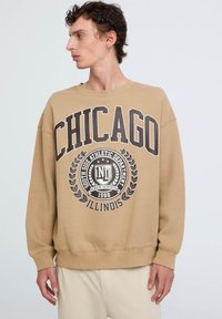 Felpa color khaki con "CHICAGO" in bold lettere nere, caratterizzata da un emblema grafico e un design di alloro. Polsini e orlo a coste.