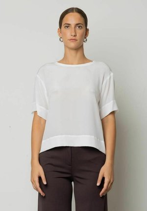 Donna che indossa una maglia bianca a maniche corte larga e pantaloni larghi scuri, in piedi davanti a uno sfondo grigio chiaro uniforme.