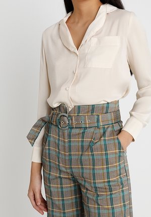Mujer que lleva una blusa crema de manga larga metida en pantalones marrones a cuadros de talle alto con un cinturón de tela a juego.