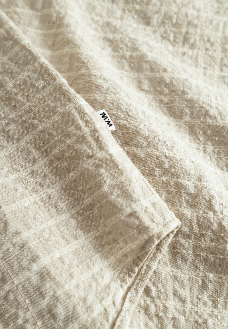 Tissu en lin beige clair présentant un motif de grille subtil. Texture douce et bords repliés avec une petite étiquette dans le coin inférieur.