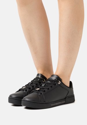 Jambes portant des baskets basses Tommy Hilfiger noires avec des lacets noirs et des breloques argentées à logo sur les lacets sur un fond blanc.