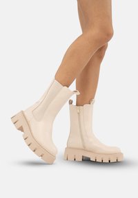 Gambe che indossano stivali alla caviglia chunky beige con piattaforma, cerniera laterale e linguette, mostrati su uno sfondo bianco uniforme.
