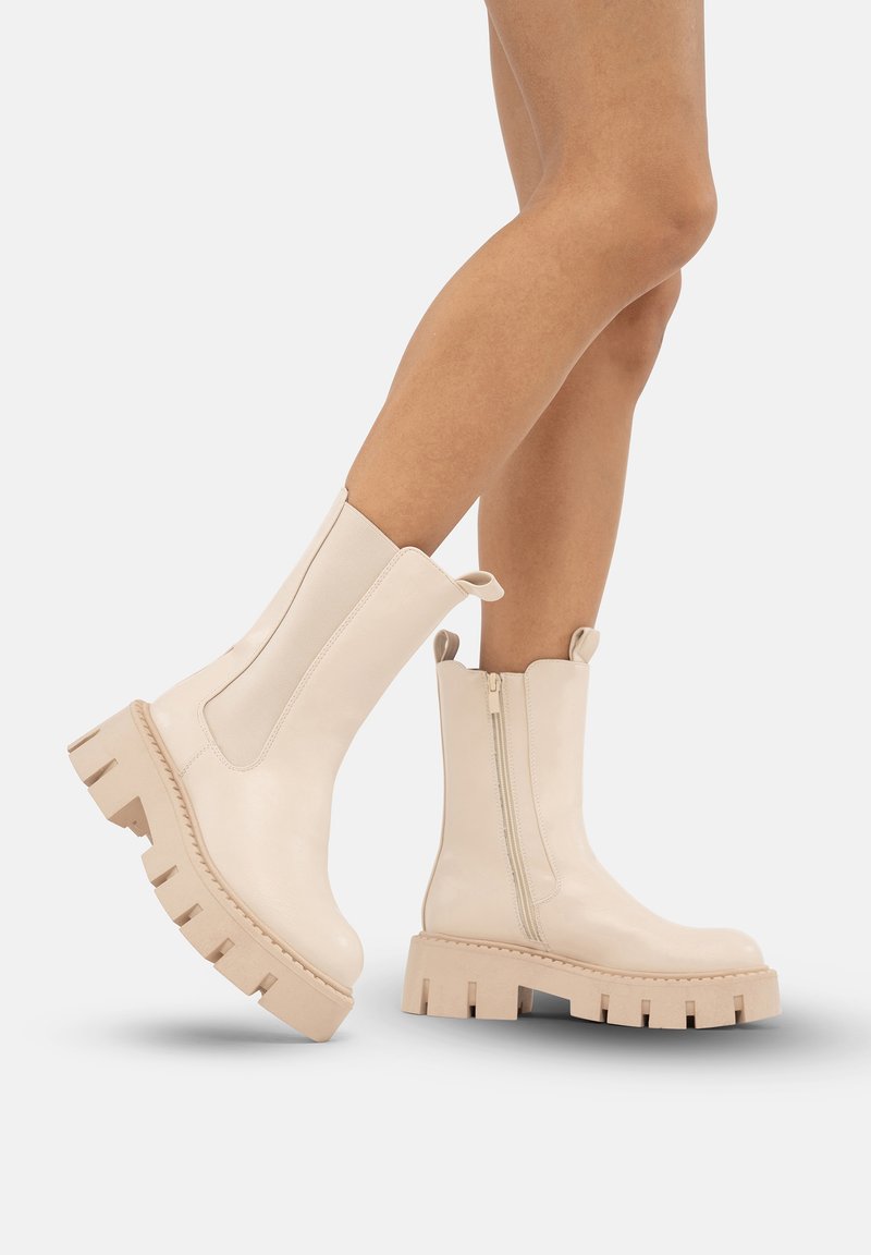 Gambe che indossano stivali alla caviglia chunky beige con piattaforma, cerniera laterale e linguette, mostrati su uno sfondo bianco uniforme.