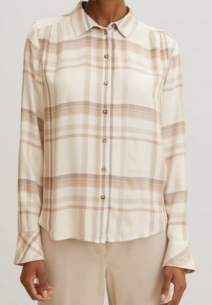 Mujer con camisa de cuadros beige y crema con botones y mangas largas, combinada con pantalones beige claros, de pie frente a un fondo liso.