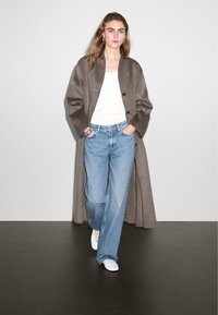 Een lange, grijze open voorkant jas over een witte tanktop, gecombineerd met lichtblauwe wijde spijkerbroek en witte sneakers. Glad wollen materiaal en eenvoudig ontwerp.
