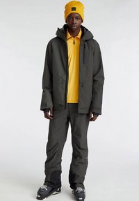 Casaco impermeável cinza escuro com capuz, camisa amarela, gorro amarelo combinando, detalhes pretos e calças de neve, usado com botas de neve cinzentas.