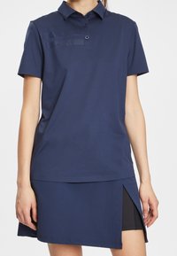 Mörkblå polo t-shirt med klassisk krage, korta ärmar och knäppning; har en perforerad randdesign på bröstet; matchas med en kjol i samma färg.