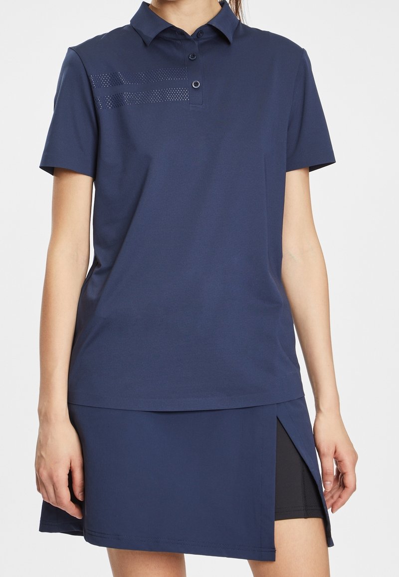 Mörkblå polo t-shirt med klassisk krage, korta ärmar och knäppning; har en perforerad randdesign på bröstet; matchas med en kjol i samma färg.