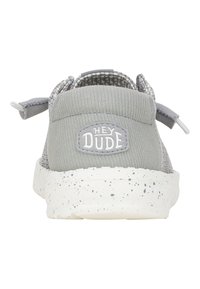Zapatos de lona gris tipo slip-on con una superficie texturizada, que cuentan con una suela salpicada de blanco y un logo circular de "HEY DUDE" en el talón.
