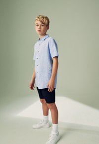 Lyseblå kortærmet skjorte med knapper, marineblå shorts, hvide sneakers og hvide knæstrømper, stående mod en sart grøn baggrund.