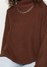 Suéter de cuello alto de punto marrón oscuro con textura de canalé, corte relajado y detalle de cadena plateada, usado sobre jeans de color azul claro.