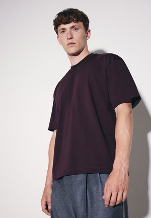 Junger Mann mit kurzen lockigen Haaren, der ein dunkelviolettes übergroßes T-Shirt und graue Hose trägt, steht vor einem einfarbigen hellen Hintergrund.