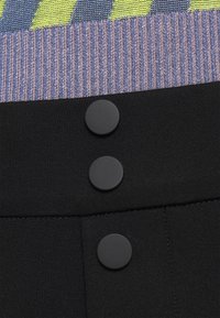 Detailní záběr na černou látku se třemi matnými černými kulatými knoflíky svisle zarovnanými pod žebrovaným pleteným materiálem v purpurových a žlutých pruzích.