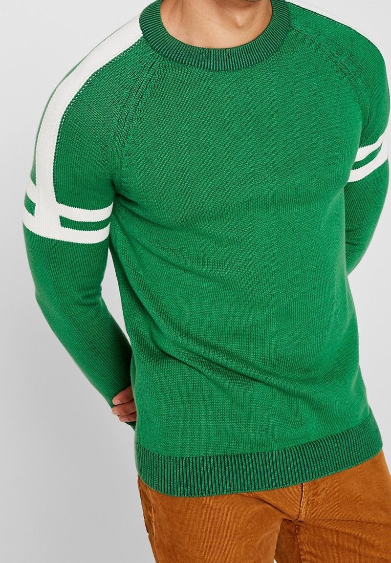 Homme portant un pull en maille vert avec des rayures blanches sur les manches et un pantalon en velours côtelé marron, debout les mains derrière le dos.