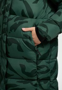 Chaqueta tipo puffer de camuflaje verde con un diseño acolchado, que cuenta con un bolsillo lateral y un acento en los puños de color negro. Fabricada con un material suave y brillante.