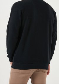 Maglione nero visto da dietro, con collo rotondo, maniche lunghe e orlo a costine. Il tessuto appare morbido e strutturato.