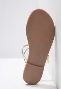Gyllene sandal med en platt, brun gummisula. Har tunna, korsande remmar och en cirkulär storleksindikator (37) på sulan.