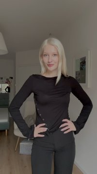 Svart langermet topp med rynket stoff og dekorative knapper ned på den ene siden, kombinert med svarte tettsittende bukser. Jevn tekstur, moderne design.