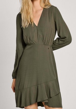 Femme portant une robe portefeuille à manches longues vert olive avec un motif texturé subtil et un ourlet asymétrique à volants.