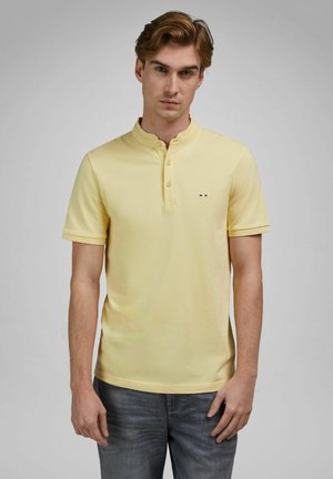 Jonge man met lichtbruin haar, gekleed in een lichtgele korte mouw henley en verweerde blauwe spijkerbroek, tegen een effen achtergrond.