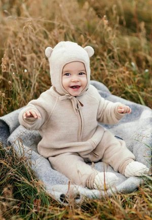 Beige babyoutfit med blødt stof, der har lynlås og bamseører på hætten. Barnet sidder på et gråt tæppe i græsset.