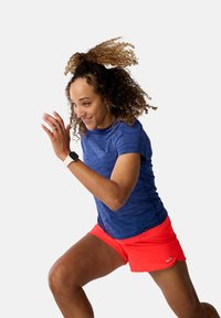 Atletka w niebieskiej koszulce sportowej i jasnoczerwonych szortach, biegnąca z kręconymi włosami w ruchu. Na nadgarstku ma smartwatcha.
