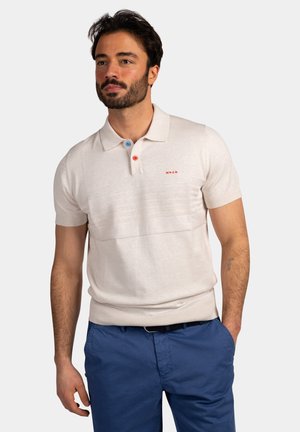Man met baard die een beige poloshirt met korte mouwen draagt met gekleurde knopen en een blauwe broek, staand met één hand in de zak.