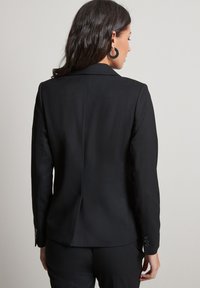 Sort skrædderjakke med en glat overflade, enkelt bagventil og knapmanchetter. Har et tætsiddende design med en struktureret silhuet.