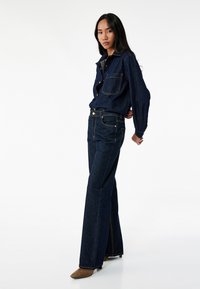 Camisa de denim y pantalones acampanados en azul oscuro con costuras doradas, con bolsillos frontales y cierre de botones. Calzado marrón con tacón.