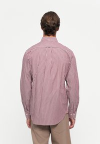 Man in een rood-wit gestreepte lange mouwen shirt en beige broek, met de rug naar de camera gekeerd tegen een effen witte achtergrond.