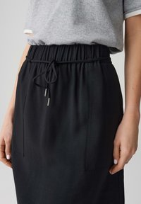 Taille und Hände einer Person, die ein graues kurzärmliges Oberteil trägt, das in einen schwarzen Rock mit elastischem Bund und Kordelzug gesteckt ist.